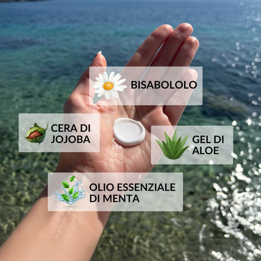Mano che mostra la texture di una crema fluida doposole con i suoi ingredienti principali: bisabololo, cera di jojoba, gel di aloe e olio essenziale di menta, su uno sfondo di mare. 
