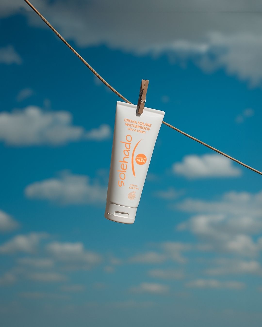 Tubo di crema solare waterproof Solehado appeso con una molletta a un filo per il bucato, sullo sfondo di un cielo azzurro con nuvole.