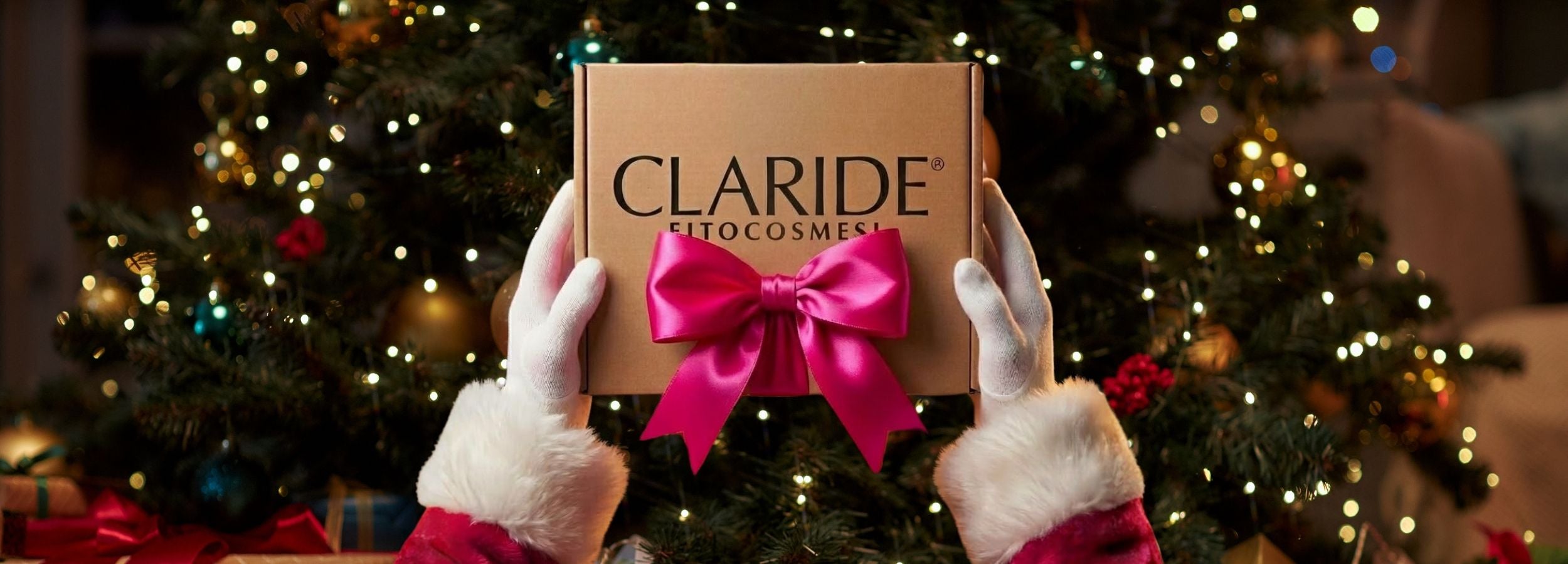 Mani di Babbo Natale con guanti bianchi e polsini di pelliccia rossa tengono una scatola regalo marrone con il logo 'CLARIDE FITOCOSMESI' e un grande fiocco di raso rosa shocking, sullo sfondo un albero di Natale illuminato e un camino con decorazioni festive sfocate.